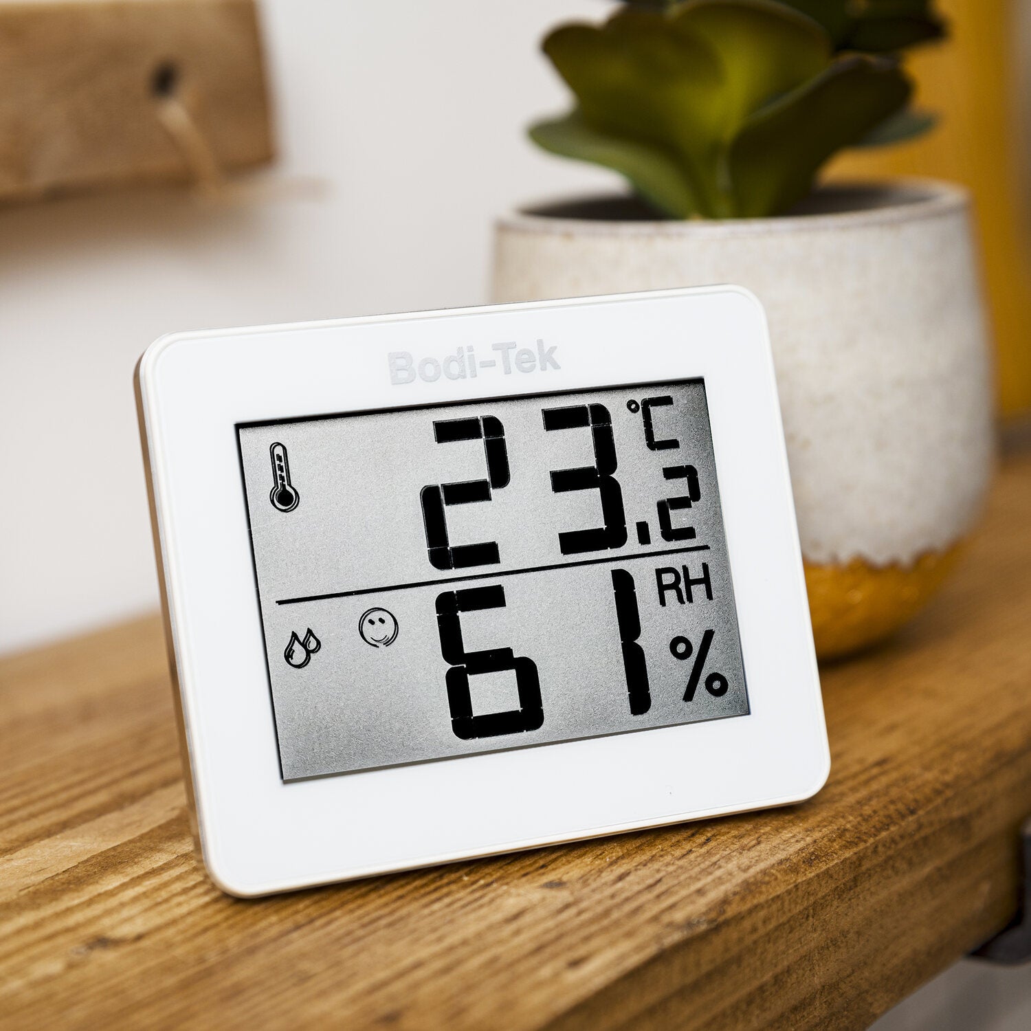 The Digital Room Thermometer and Hygrometer shows 23°C and 61% humidity on a wooden surface beside a potted plant.