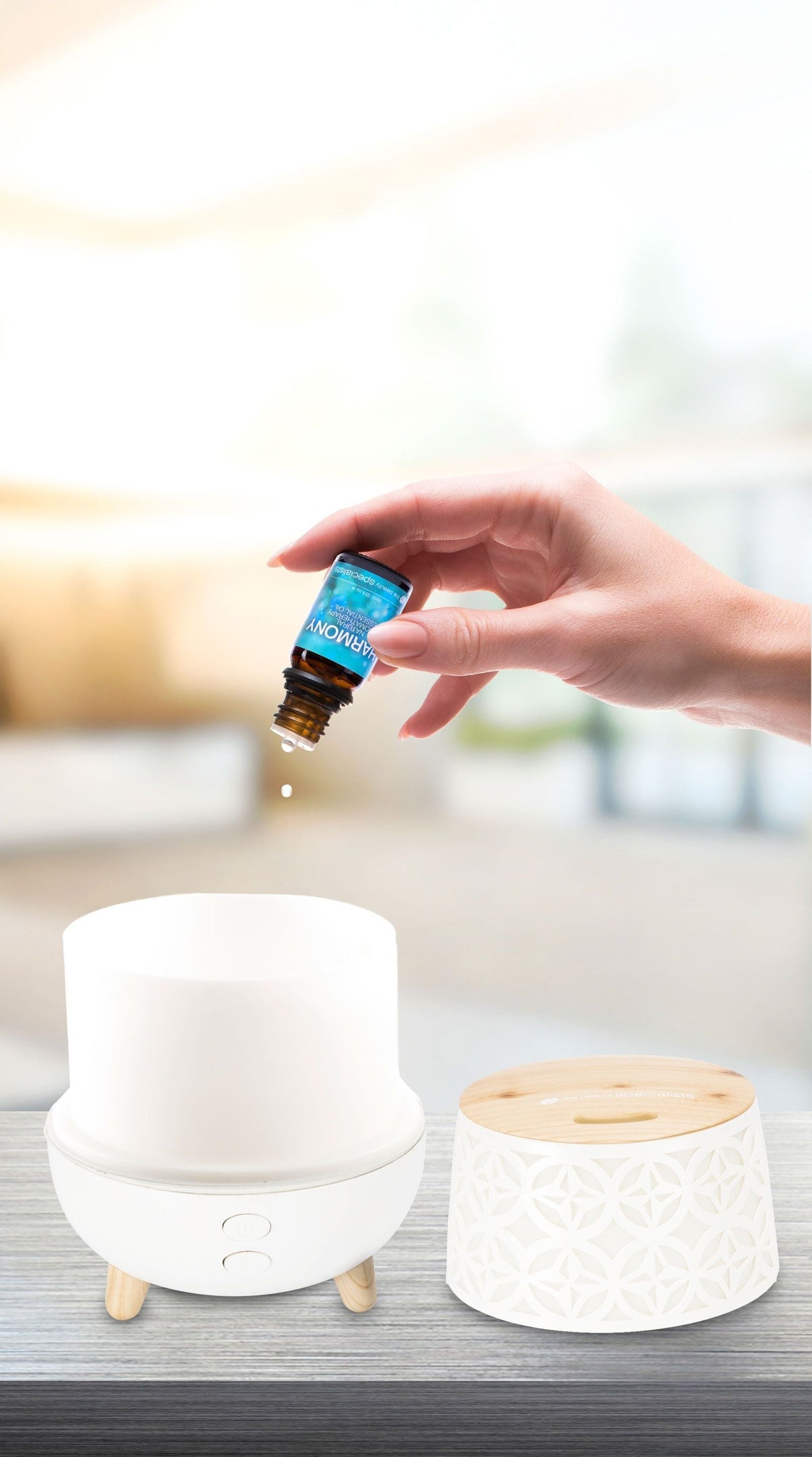 A hand pours Rio Wellbeing Collection Aromatherapy Essential Oils from a small bottle into a white electric diffuser on a table, with the decorative lid set nearby and a softly blurred background.