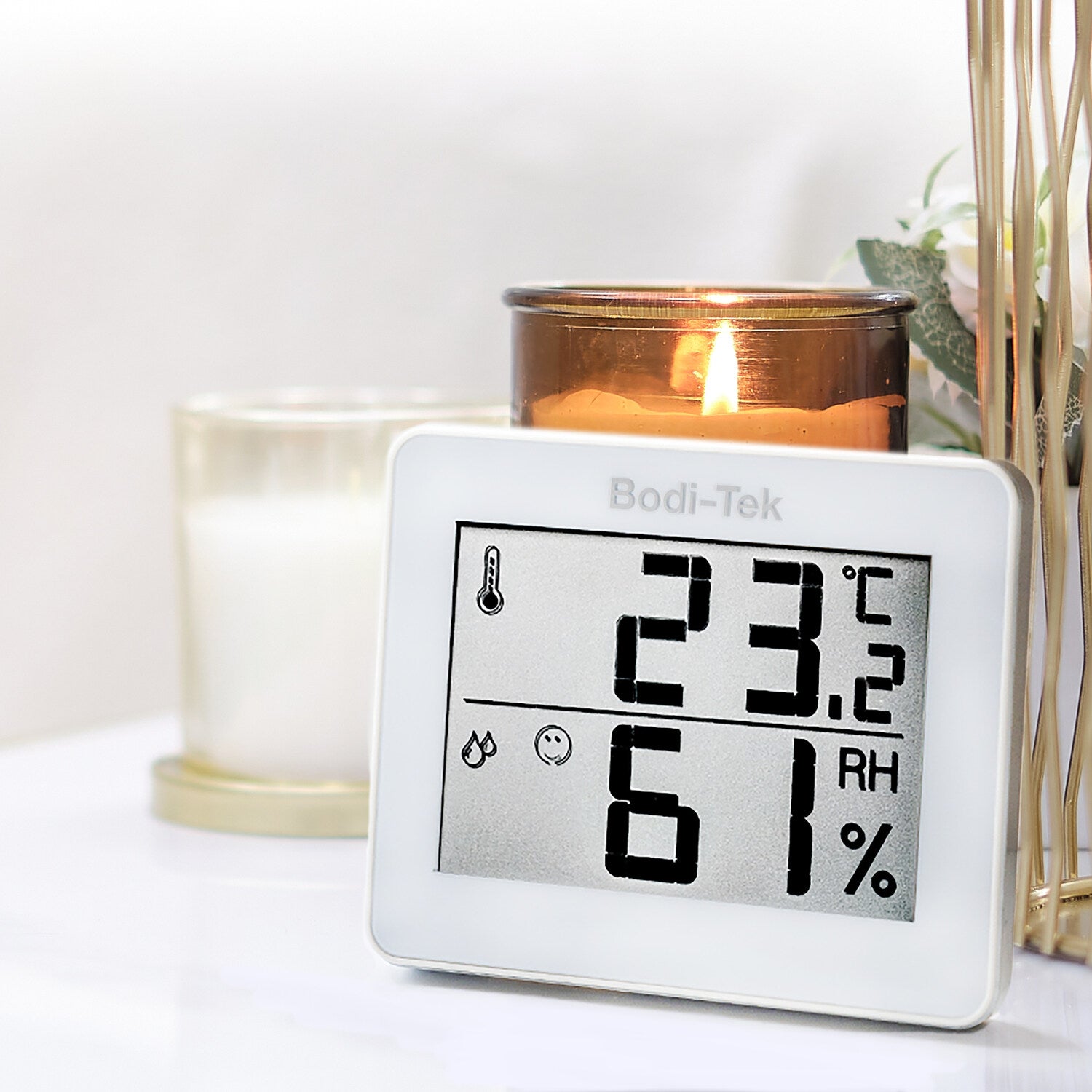A Digital Room Thermometer and Hygrometer showing 23°C and 61% humidity rests on a white surface beside both lit and unlit candles, with a small floral arrangement in the background.