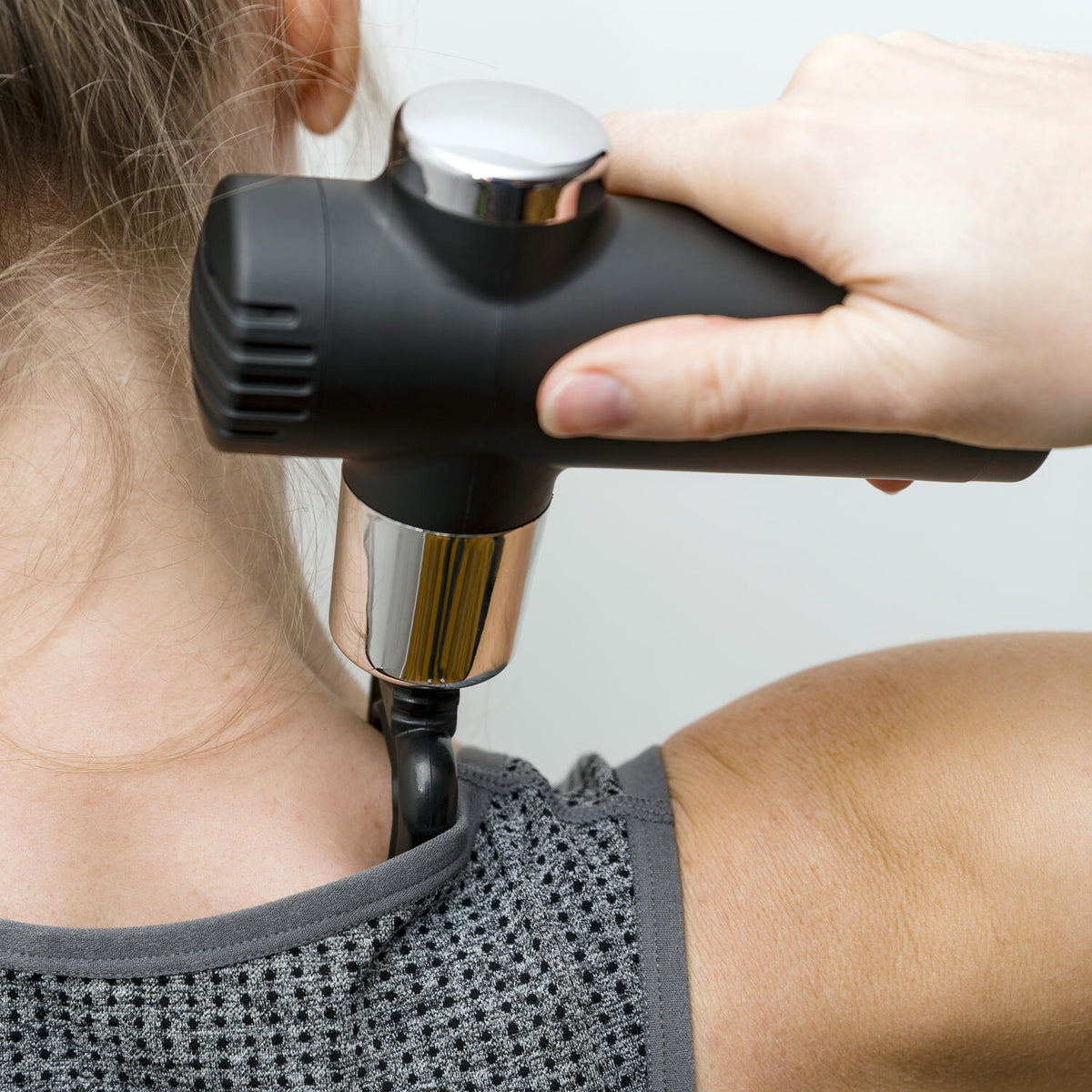 A person uses a Mini Deep Tissue Massage Gun on their neck and shoulder while wearing a sleeveless gray textured top, holding the portable massager in their right hand.