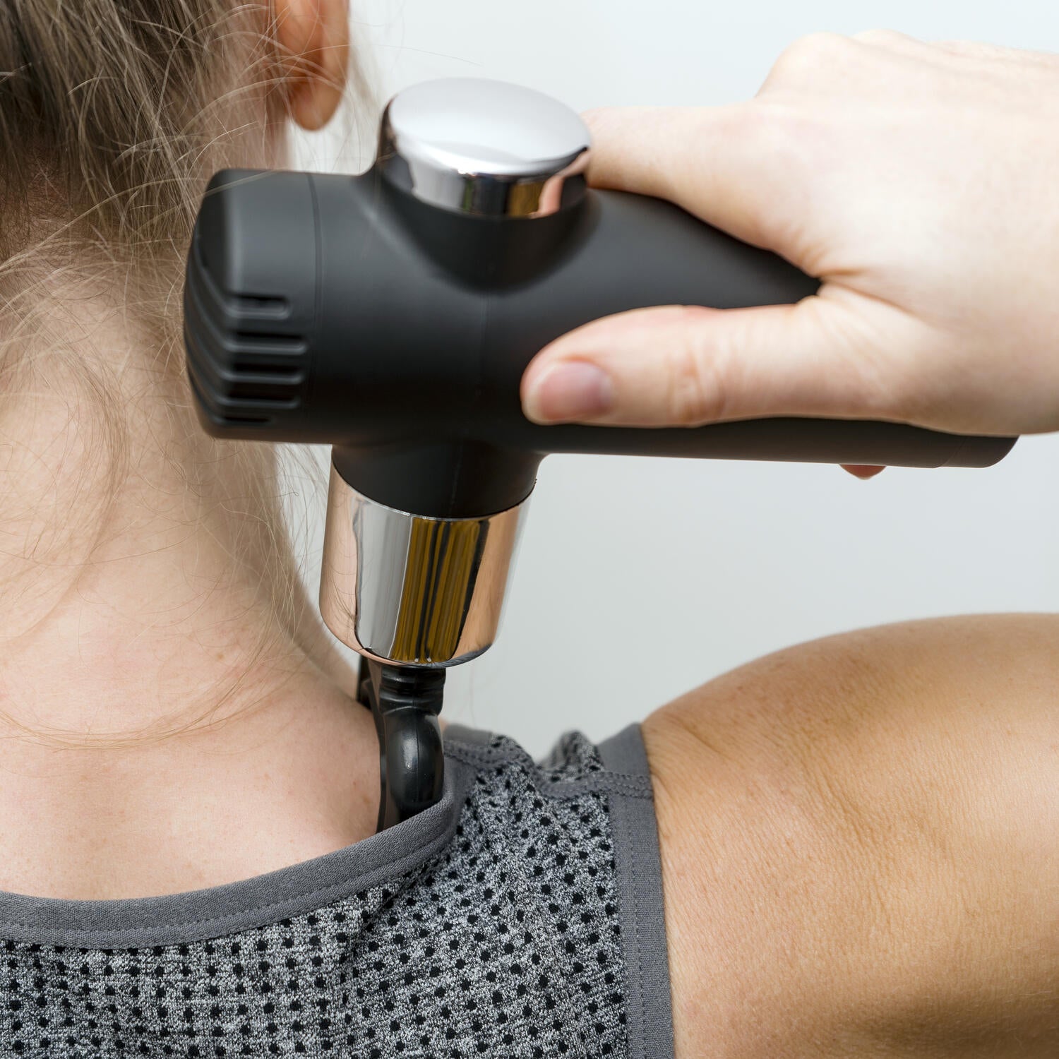 A person uses a Mini Deep Tissue Massage Gun on their neck and shoulder while wearing a sleeveless gray textured top, holding the portable massager in their right hand.