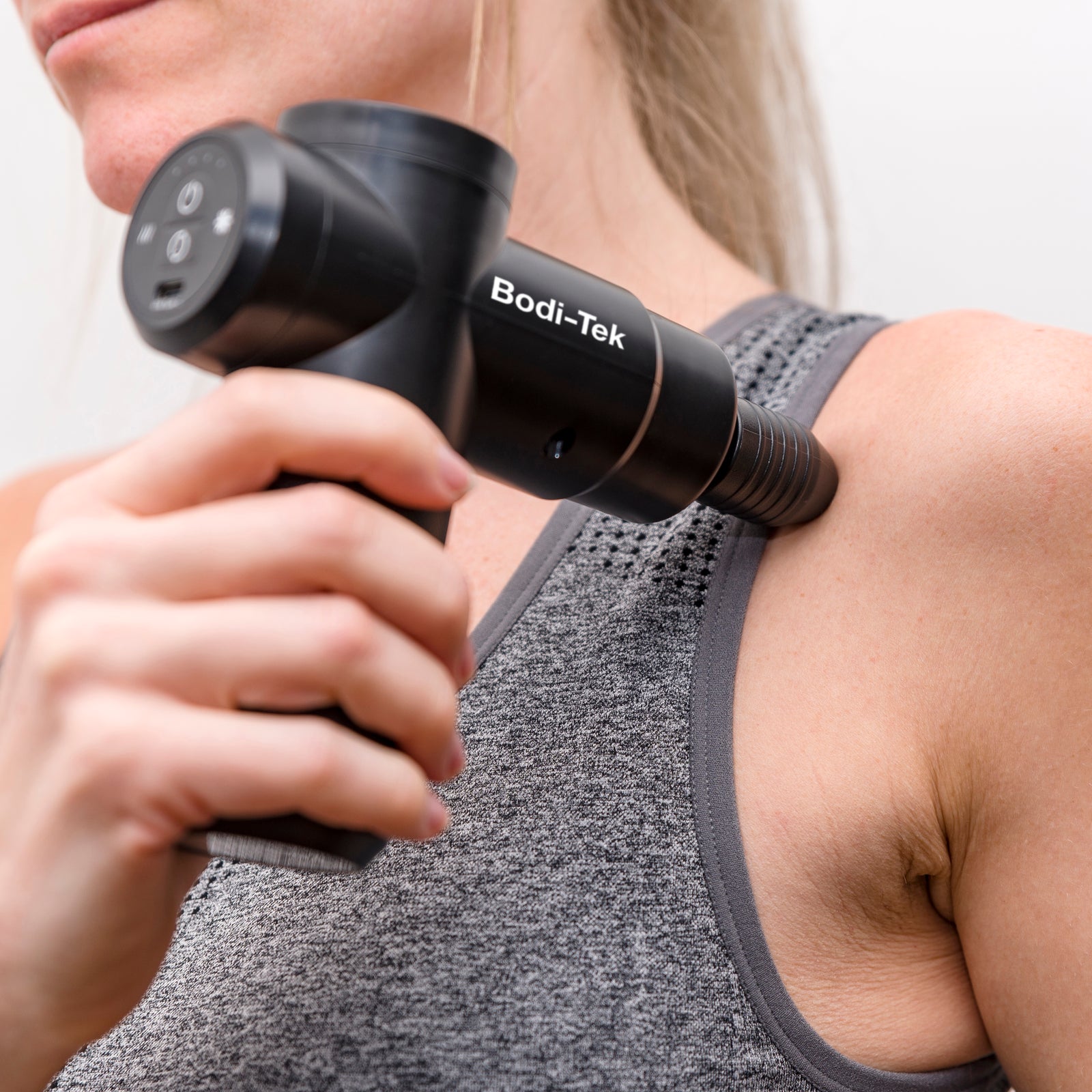 A person uses the Hot & Cold Deep Tissue Massager Gun in black on their upper shoulder near the collarbone while wearing a grey athletic tank top, targeting the area for effective muscle recovery.