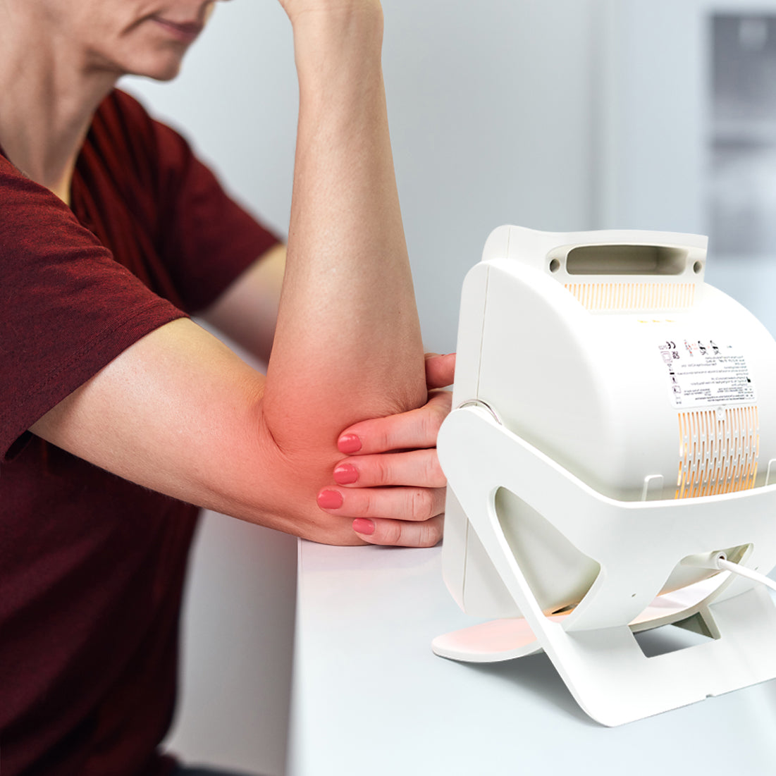 A person in a red shirt sits at a table, exposing their reddened forearm to an Infrared Heat Lamp, likely using it for pain relief or to promote blood circulation.