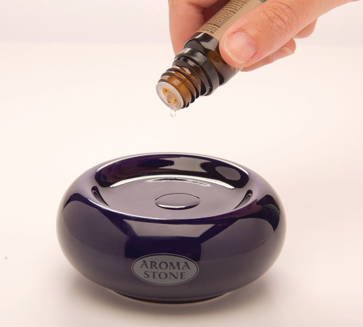 A hand pours essential oil from a small bottle onto an Aroma Stone, a natural black ceramic oil diffuser, against a plain background.