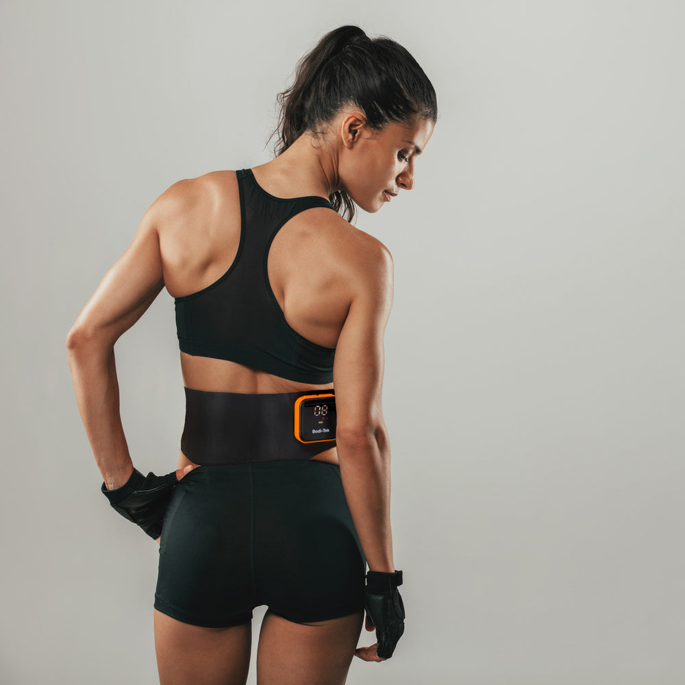 A fit woman in black athletic wear and gloves stands with her back to the camera, displaying the Ab & Back Muscle Strengthener with heat strapped around her lower back against a plain light gray background.