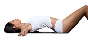 A woman in white athletic wear lies on her back, arms outstretched, using The Original BackMagic with Thermal Gel Heat Pads for lower back support and pain relief against a plain white background.