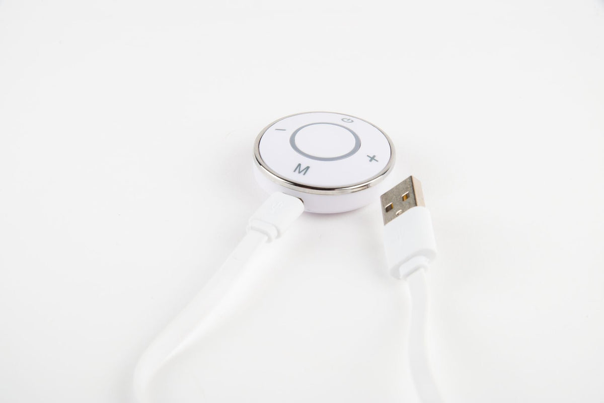 A close-up of the Circulation Plus Boost & Revive Mat, a white portable leg exerciser with a circular control panel featuring plus, minus, M, and power buttons, connected to a white USB cable on a white background.